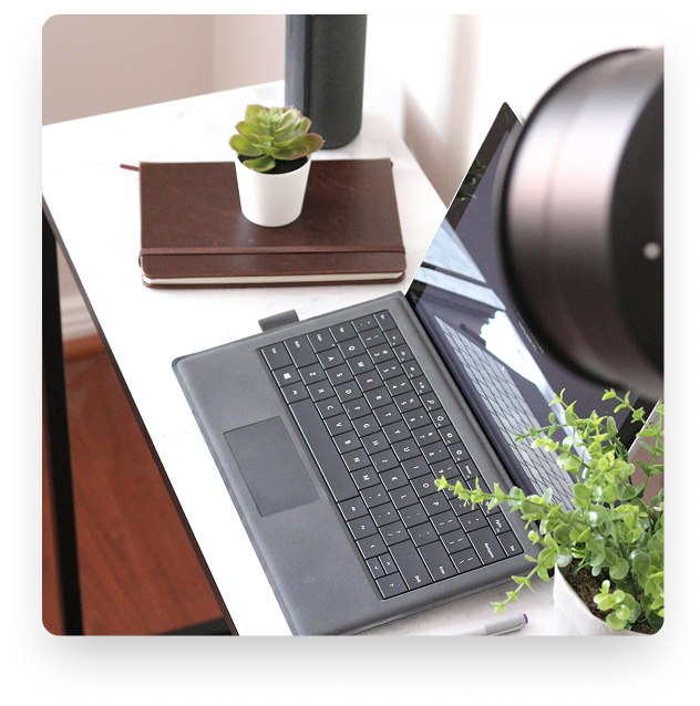 Un ordinateur portable noir est posé sur un bureau blanc, entouré de plantes vertes et d'un carnet en cuir marron à côté d'un petit pot de plante succulente.